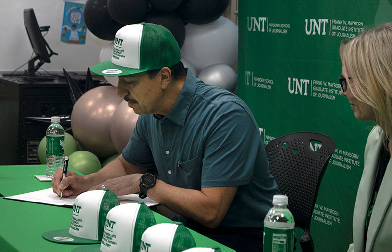 George Esquivel sits in front of a UNT Mayborn School of Journalism backdrop at a signing event